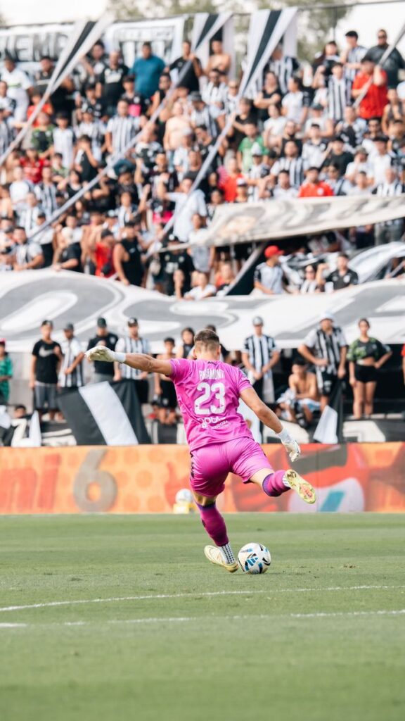 Goalkeeper in pink uniform kicks ball during a lively soccer match with cheering crowd.